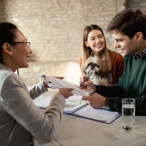 Young happy couple signing document while having meeting with real estate agent in the office. Focus is on man.